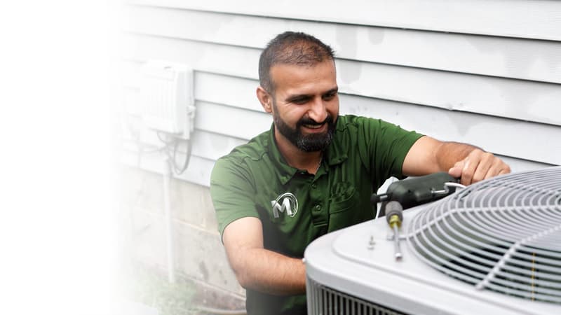 Smiling HVAC technician in a green uniform using a tool to service an outdoor air conditioner condenser beside a home